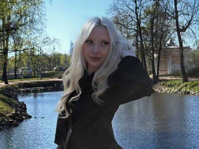 MargretMurak with long, wavy white hair, wearing a black shirt, standing by a river, hand in hair, looking slightly to the side with a subtle smile.