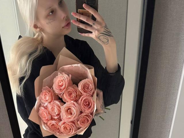 MargretMurak with pale blonde hair in ponytail, holding pink rose bouquet, wearing black shirt, tattoo on left forearm, taking mirror selfie, holding phone, pink nails, neutral expression.