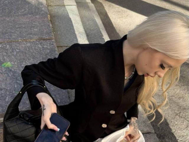 MargretMurak with long blonde hair, black blazer, holding black bag and phone, looking down at white package, tattoo on right hand, standing on stone sidewalk.
