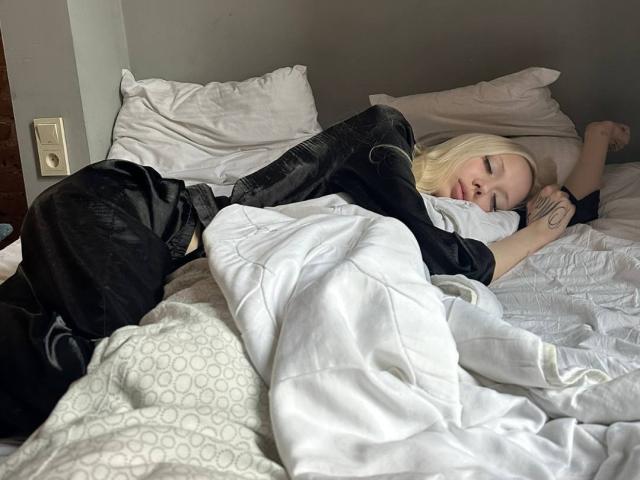 MargretMurak is lying in bed, asleep with black pants on, white quilt covering her ass, blonde hair, tattoo on her right arm, and gray walls in the background.