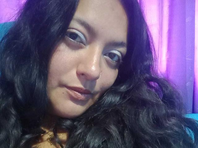 FabritziaPiel has wavy black hair, a nose ring, and light makeup with blue eyeshadow. She's looking slightly to the side with a subtle smile, showing her brown lips.