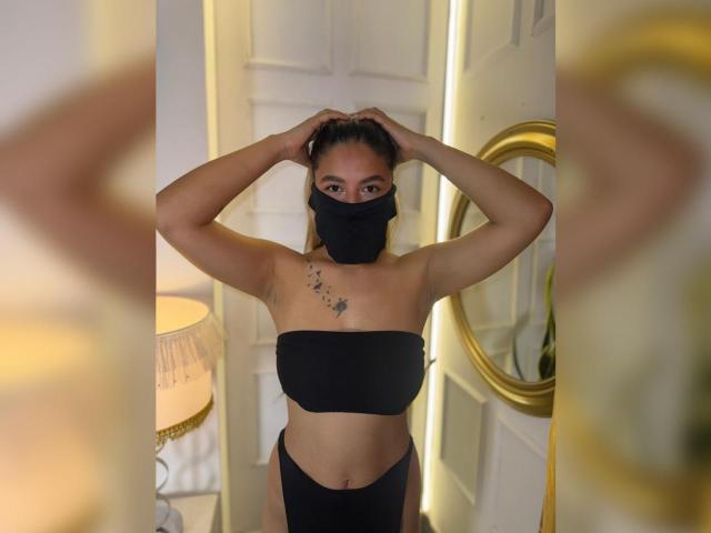 AlanaCuttie standing with arms raised, wearing a black mask, strapless black top, and black high-waisted underwear, showing her tits and small bird tattoo on chest.