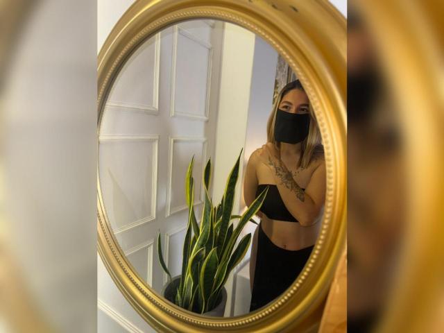 AlanaCuttie's reflection in a gold mirror, wearing a black mask, black bra, and black pants, with a green snake plant in the background, showing tattoos on her chest.