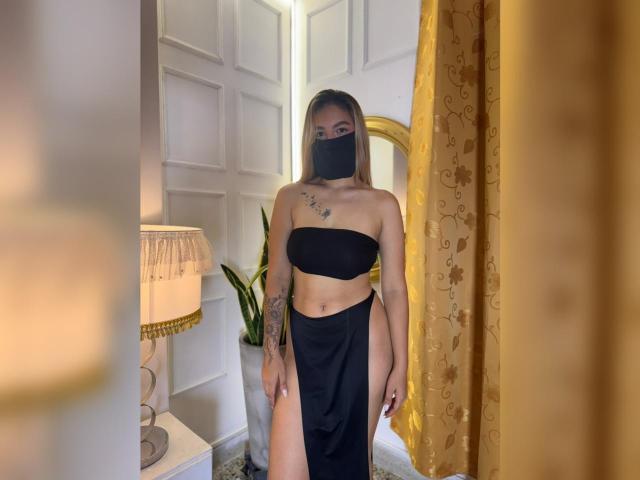 AlanaCuttie stands in a room wearing a black tube top and high-waisted skirt, black mask covering her mouth, tattoos on her right arm, showing her ass.
