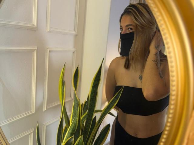AlanaCuttie with black mask, black strapless top, black pants, blonde hair, tattoo on right arm, standing by mirror, green plant, white door, golden mirror frame.
