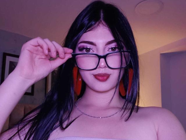 IsabellaaRossi with dark hair, red hoop earrings, black glasses, red lipstick, and a silver necklace, holding glasses, showing off her tits in a strapless top.