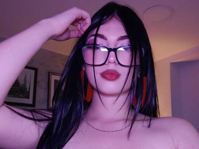 IsabellaaRossi with long black hair, glasses, red earrings, and red lipstick, touching her hair, showing bare shoulders, wearing a silver necklace, in a purple-lit room.