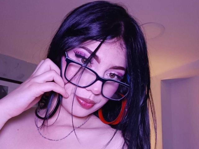 IsabellaaRossi with black hair, glasses, and red headphones, holding her glasses, showing her tits, pink eyeshadow, and a subtle smile, in a purple-lit room.