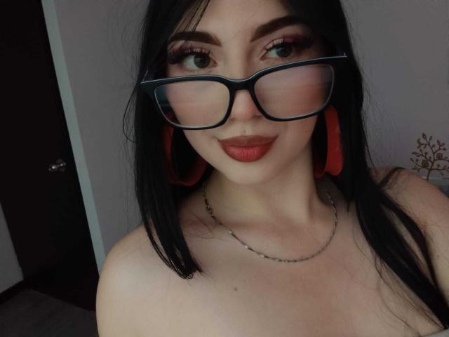 IsabellaaRossi with black hair, glasses, red earrings, and red lipstick, showing off her tits, smiling at the camera, wearing a chain necklace.