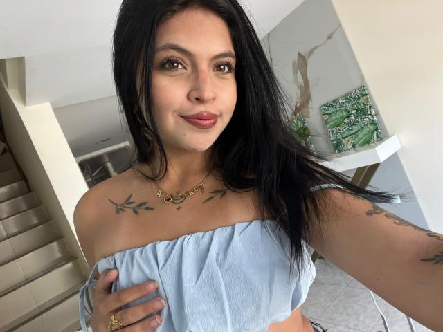 NyraVanel with long black hair, light skin, small tattoos on chest and arms, wearing a light blue off-shoulder top, smiling, holding top with left hand, gold necklace.