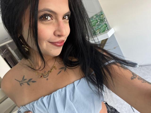 NyraVanel with long black hair, light blue off-shoulder top, small tits, tattoos on shoulders and arm, pink lipstick, gold necklace, smiling at camera, white background, indoor setting.