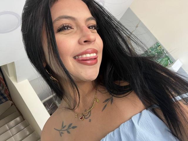 NyraVanel, con cabello negro largo, sonriendo, vestida con una blusa azul claro con un hombro descubierto, mostrando tatuajes en el cuello y el pecho, y un collar de oro con una "M".".