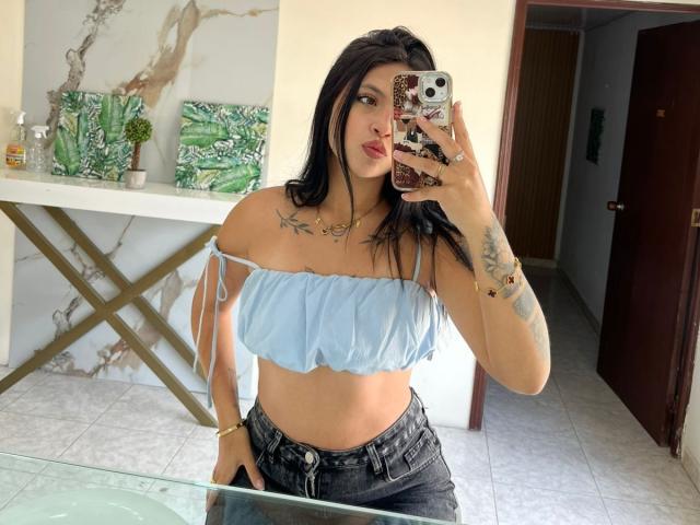 NyraVanel with black hair, blue off-shoulder top, tattoos on chest and arm, wearing black jeans, holding phone with floral case, pouting, standing in front of mirror.