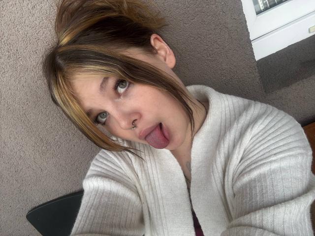 EthelynWiskowski with messy brown and blonde hair, green eyes, nose ring, and tongue out, wearing a white ribbed sweater, looking up at the camera.
