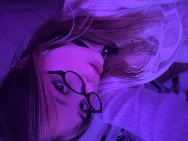 EthelynWiskowski's face with purple lighting, wearing glasses, black choker, and white shirt, lying down with long brown hair, nose ring, and tattoo on her right cheek.