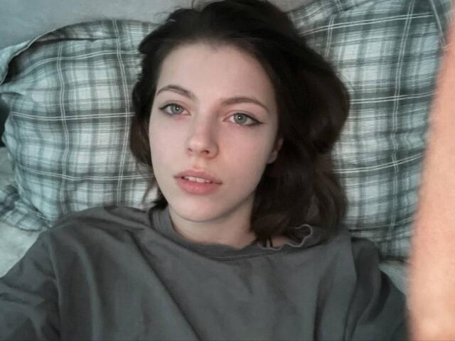 JennellMatacale lying in bed, wearing a gray shirt, with wavy brown hair, green eyes, and slightly parted pink lips, looking up with a soft expression.