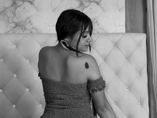 SamanthhaLove, with her back to the camera, sits on a tufted bed, wearing a strapless, ruffled dress, showing a tattoo on her left shoulder and a necklace.