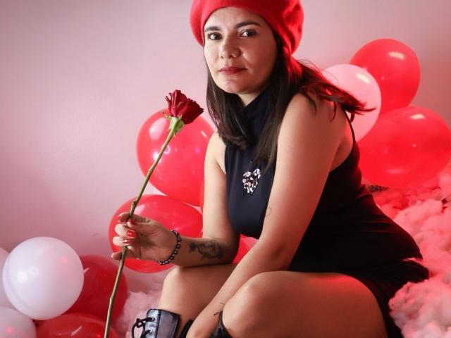 BulmaHot with red beanie, black dress, holding red rose, red and white balloons, and black boots, sitting on fluffy pink, showing tattoo on right forearm, looking serious.