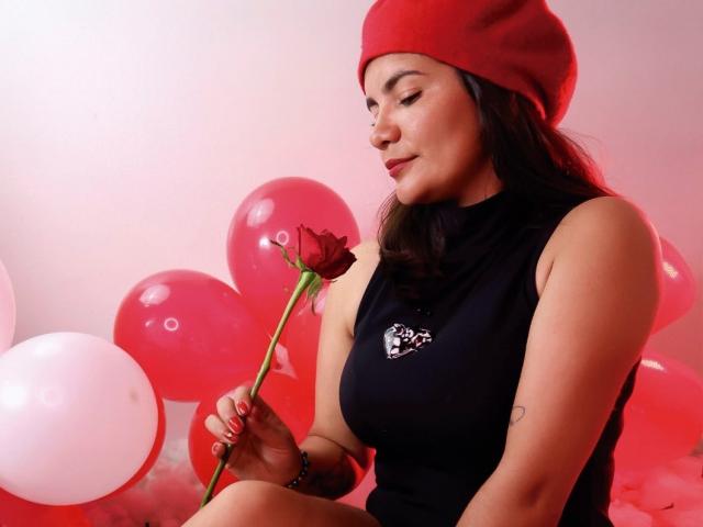BulmaHot with red beanie, black sleeveless top, holding red rose, sitting with red and white balloons, eyes closed, soft smile, holding rose stem with red nail polish.