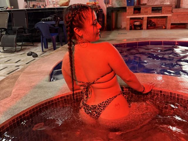 LorensDouce in a leopard print bikini, wet red hair in a braid, sitting in a hot tub, back to camera, showing her ass and small tits, water glowing red.