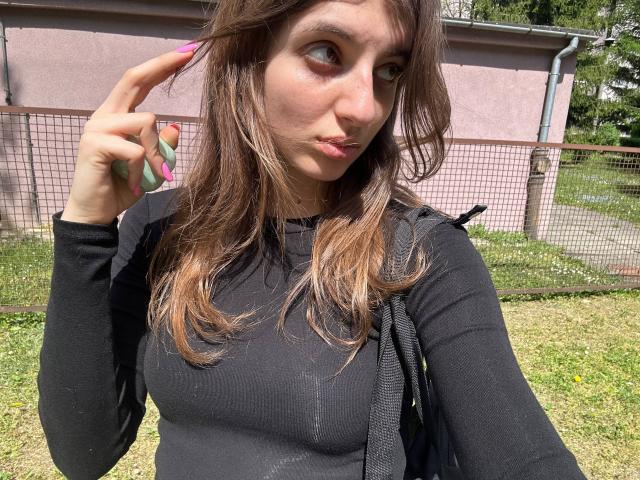 KarieMestler with long brown hair, wearing a black top, showing a peace sign with a green and pink fingernail, small tits, and a black backpack.