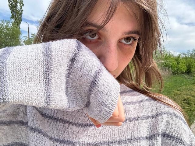 KarieMestler with dirty brown hair, green eyes, and freckles, wearing a striped gray sweater, covering her nose and mouth with a white knit sweater, outdoors.