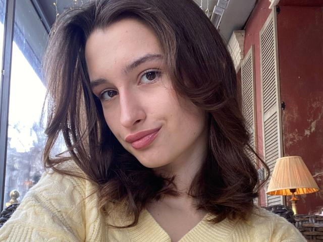 CharleenTilton with wavy brown hair, light pink lips, and a slight smile, wearing a yellow sweater, sitting near a lamp in a warmly lit room.
