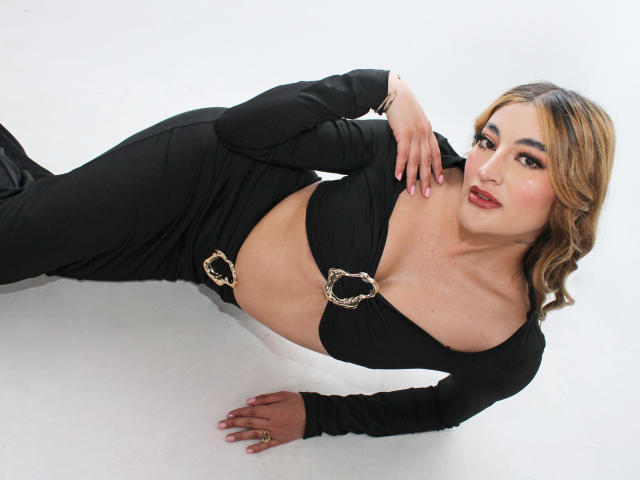 ArianaCarsson is laying down in black clothes with a cutout top, showing her tits, and gold rings. She has wavy blonde hair, red lipstick, and a cute smile.