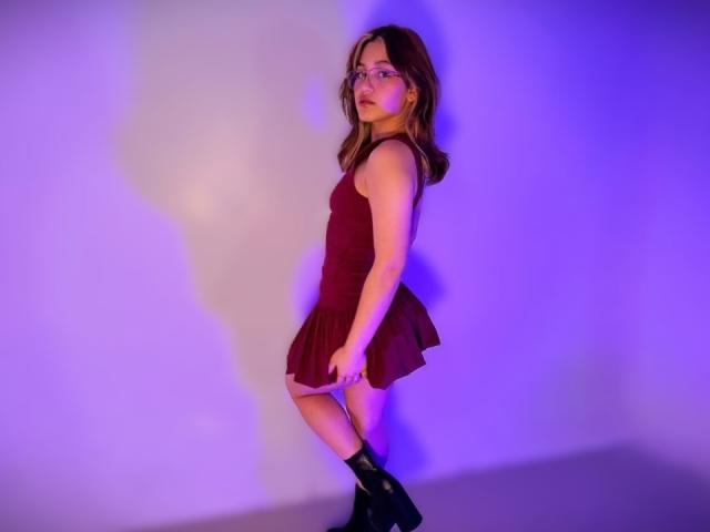 ElodieGardner with glasses, brown dress, flirty skirt, black boots, standing sideways, looking over shoulder, purple-blue lighting, shadow on wall, brown hair, cute, sexy.