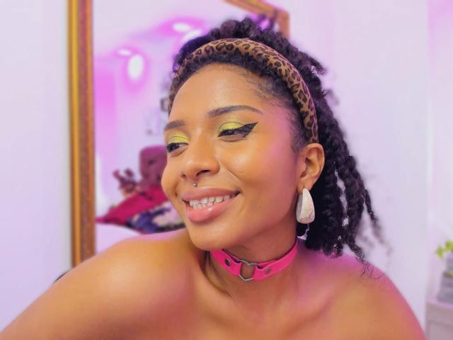 MeganWilliams with dark curly hair, leopard print headband, neon green eyeshadow, black winged eyeliner, pink choker, white earrings, smiling, nose ring, topless, soft pink and white lighting.