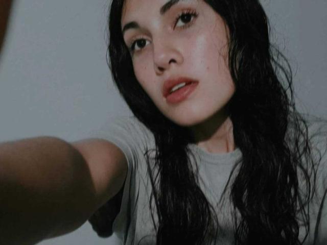 LucyGale with wet black hair, slightly parted lips, and a gray shirt, extending her arm, showing her soft, freckled face and subtle makeup in a slightly blurry selfie.