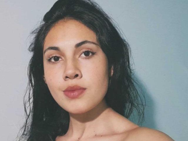 LucyGale has medium brown skin, dark wavy hair, brown eyes, and pink lips. She's topless, showing her collarbones. Her face is slightly angled to the left.