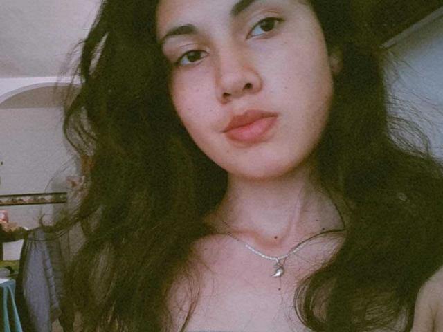 LucyGale with wavy black hair, light skin, and full lips, wearing a silver necklace with a crescent moon pendant, looking slightly to the side with a relaxed expression.