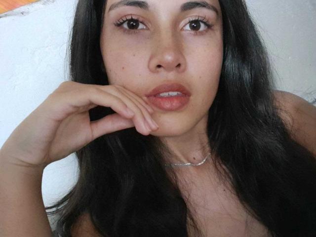 LucyGale with long black hair, brown eyes, and pinkish lips, touching her chin with her right hand, wearing a thin necklace, and showing her teeth.