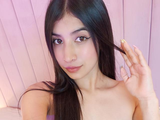 Naia Sanders, con cabello negro largo, sombra de ojos rosa, piercing en la nariz y piel pálida, sosteniendo su cabello, mostrando sus hombros desnudos y sus dedos con uñas rosas, sonriendo suavemente..