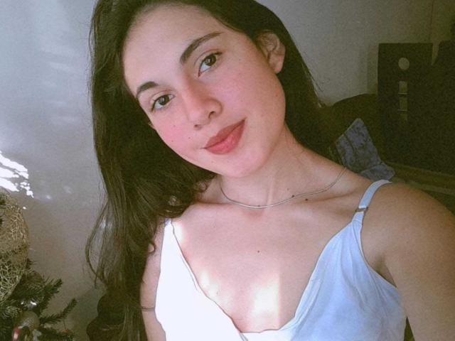 Lucy Gale, met lang donker haar, lichte huid, groene ogen en roze lippen, draagt een witte tanktop met een dunne zilveren ketting, waarbij haar kleine borsten zichtbaar zijn. Zacht zonlicht..