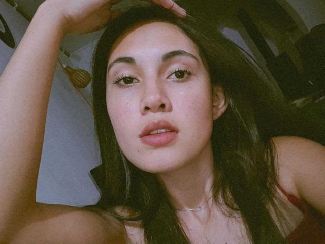 Lucy Gale, met donkerbruin haar, lichte huid en volle lippen, die lichtjes open zijn, draagt een rode top, heeft haar hand in haar haar, kijkt naar de camera, heeft subtiele make-up op, draagt een ketting en heeft een nonchalante uitdrukking..