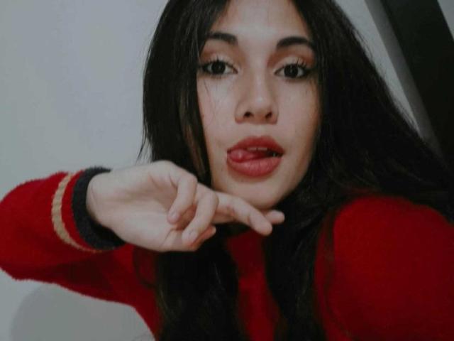 Lucy Gale, met lang zwart haar, rode lippenstift en een rode trui, wijst met een vinger naar haar lichtjes geopende mond, en kijkt verleidelijk. Haar borsten zijn niet zichtbaar..