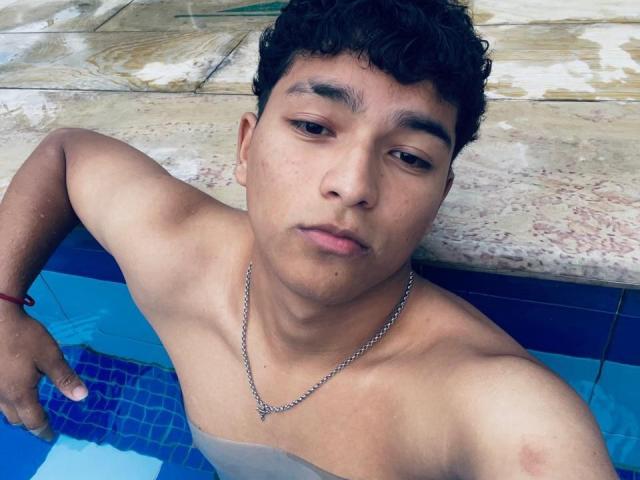 BrandonWesley, con capelli ricci e neri, pelle color nocciola, indossa una catena d'argento e un braccialetto rosso, in una piscina con piastrelle blu, guarda verso l'alto con un'espressione neutra..