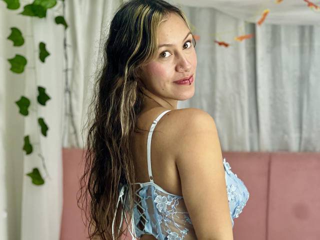 AudryLorena met lang, golvend bruin haar, een blauwe kanten BH, kijkt over haar schouder, waarbij haar borsten zichtbaar zijn, roze lippenstift, lichte huid, groene ogen, in een roze en witte kamer..