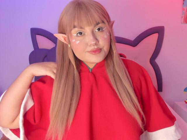 MarlaBriard with blonde elf ears, red cosplay outfit, and sparkly face, has her hand on her shoulder, showing off her pouty lips and cute nose.