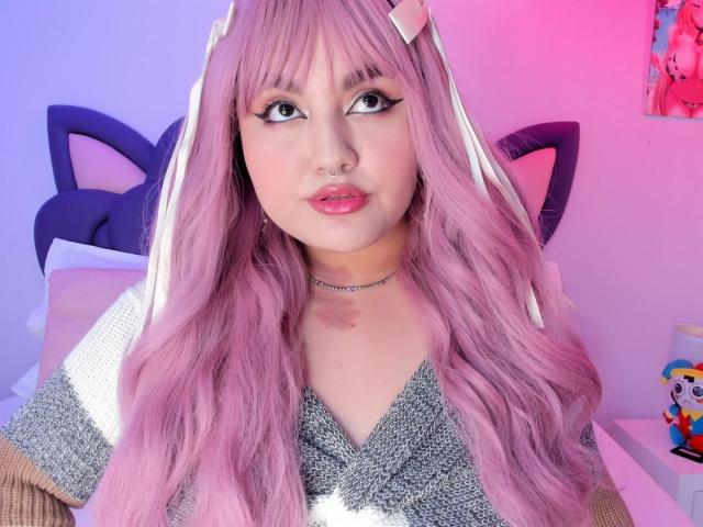 MarlaBriard with pink hair, cat ears, and white ribbons, wearing a gray and white knit top, has a nose ring and a choker, with pink and blue lighting.
