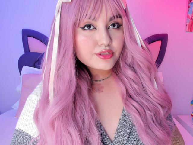 MarlaBriard with pink wavy hair, cat ears, nose ring, black eyeliner, pink lipstick, grey sweater, white ribbons in hair, and a necklace, looking at the camera.