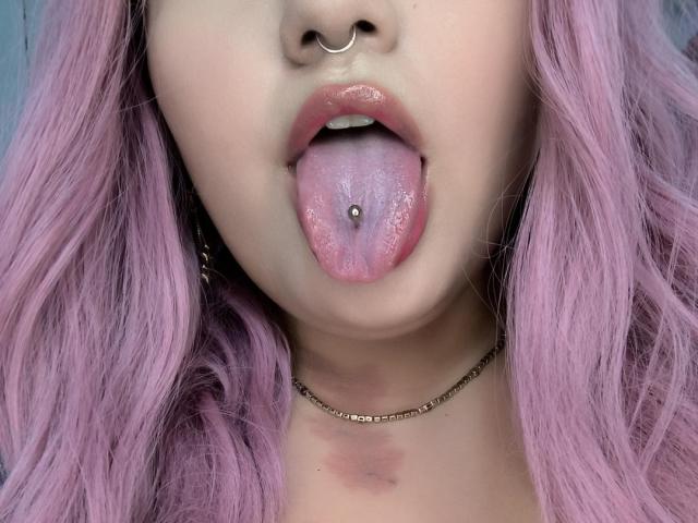 MarlaBriard with pink hair, nose ring, and tongue piercing, tongue out, showing her tongue ball. She's got a choker and a small bruise on her neck.