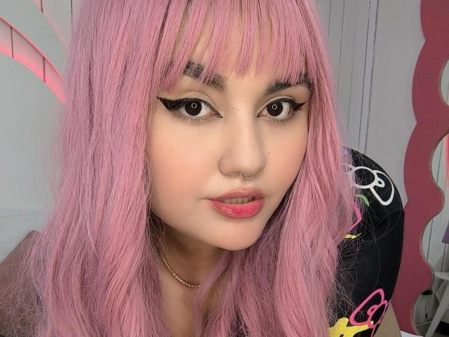 MarlaBriard with pink hair, black eyeliner, nose ring, pink lipstick, gold necklace, and cute face, looking at camera with a slight smile, showing her sexy ass.