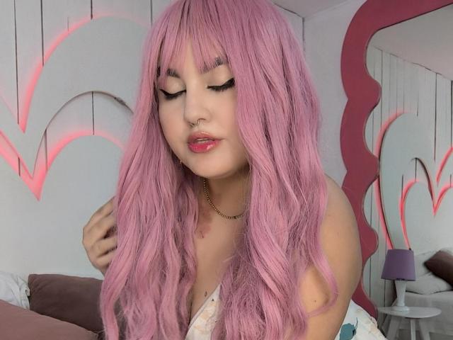 MarlaBriard with pink hair, closed eyes, black eyeliner, nose ring, and pink lipstick, holding her hair, wearing a white top, in a pink and white room.