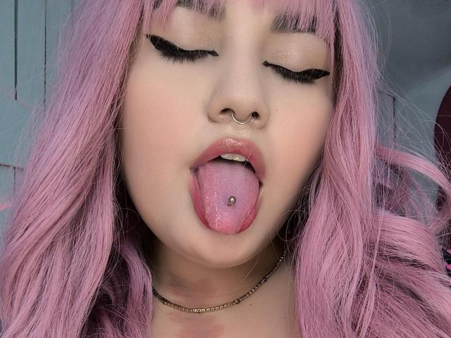 MarlaBriard with pink hair, eyes closed, nose ring, tongue out, tongue piercing, shiny pink lips, black eyeliner, gold chain, and pale skin.