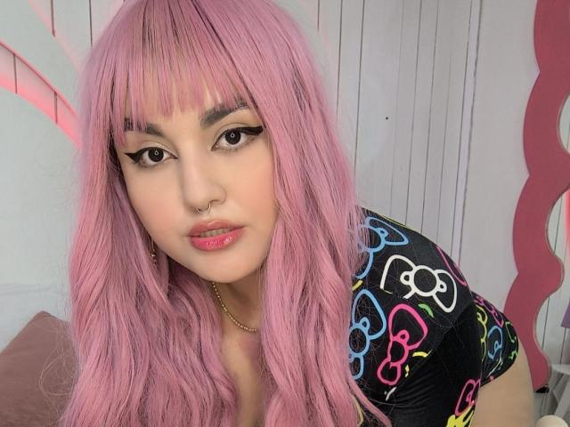MarlaBriard has pink hair, a nose ring, and eyeliner with thick lashes. She wears a colorful top and has pink lipstick, looking at the camera with a slight smile.