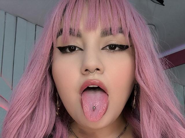 MarlaBriard has pink hair, a nose ring, and her tongue out with a tongue piercing. Her eyes are lined with black eyeliner. Her lips are slightly parted.