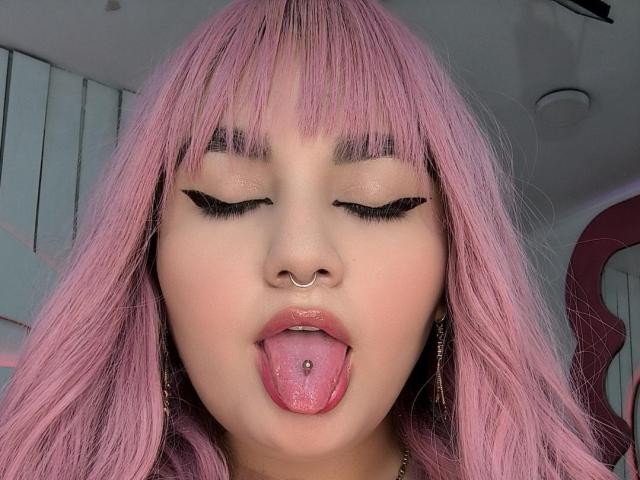 MarlaBriard has pink hair, closed eyes with black eyeliner, nose ring, and is sticking out her tongue with a tongue piercing, looking cute and sexy.